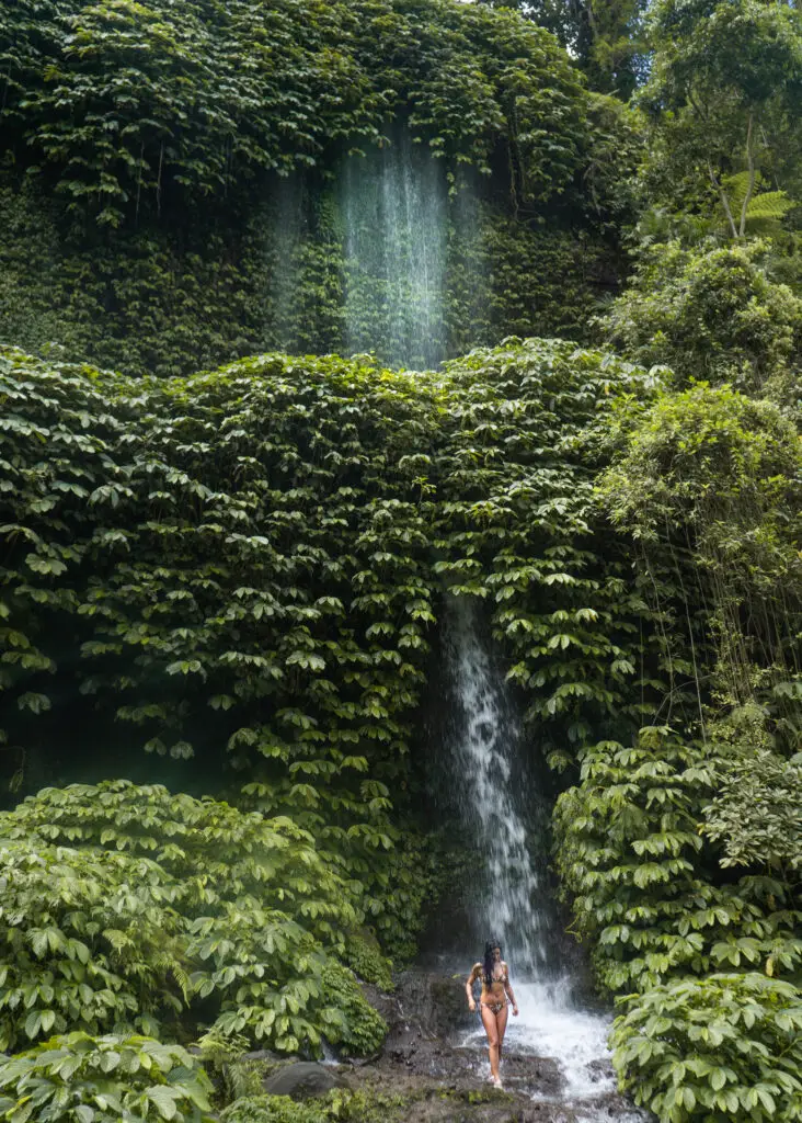 a girl in a bikini at a free falling waterfall in lombok against a wall of green leaves and trees