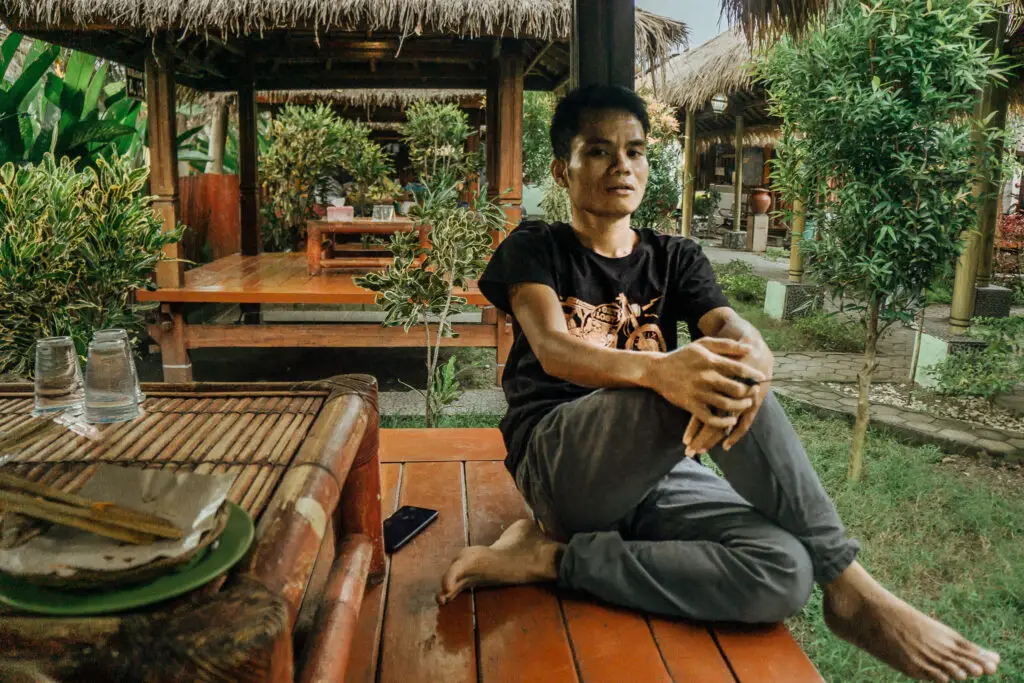 an indonesian man at an outdoor restaurant in lombok with grass huts and trees