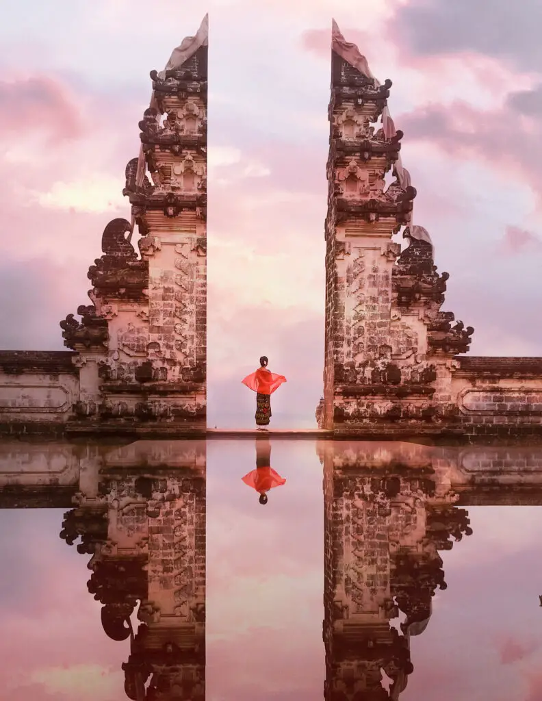 a girl in a dress during sunrise at the templ in the sky in bali with water reflecting the temple and the clouds in the background