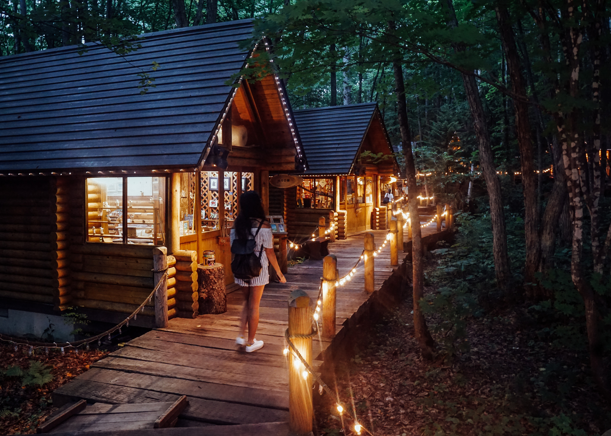 A girl walking a path next to some wooden huts lit up by fairy lights in a forest in Biei, Hokkaido