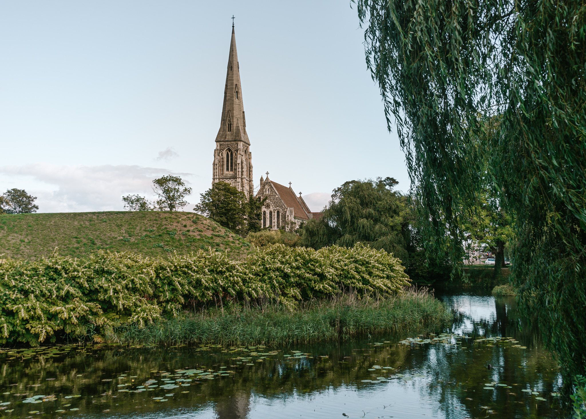 St. Albans Church with a tall spire coming from an old stone church in a grassy area on a hill, overlooking a green pond with lily pads in Kastellet, Copenhagen