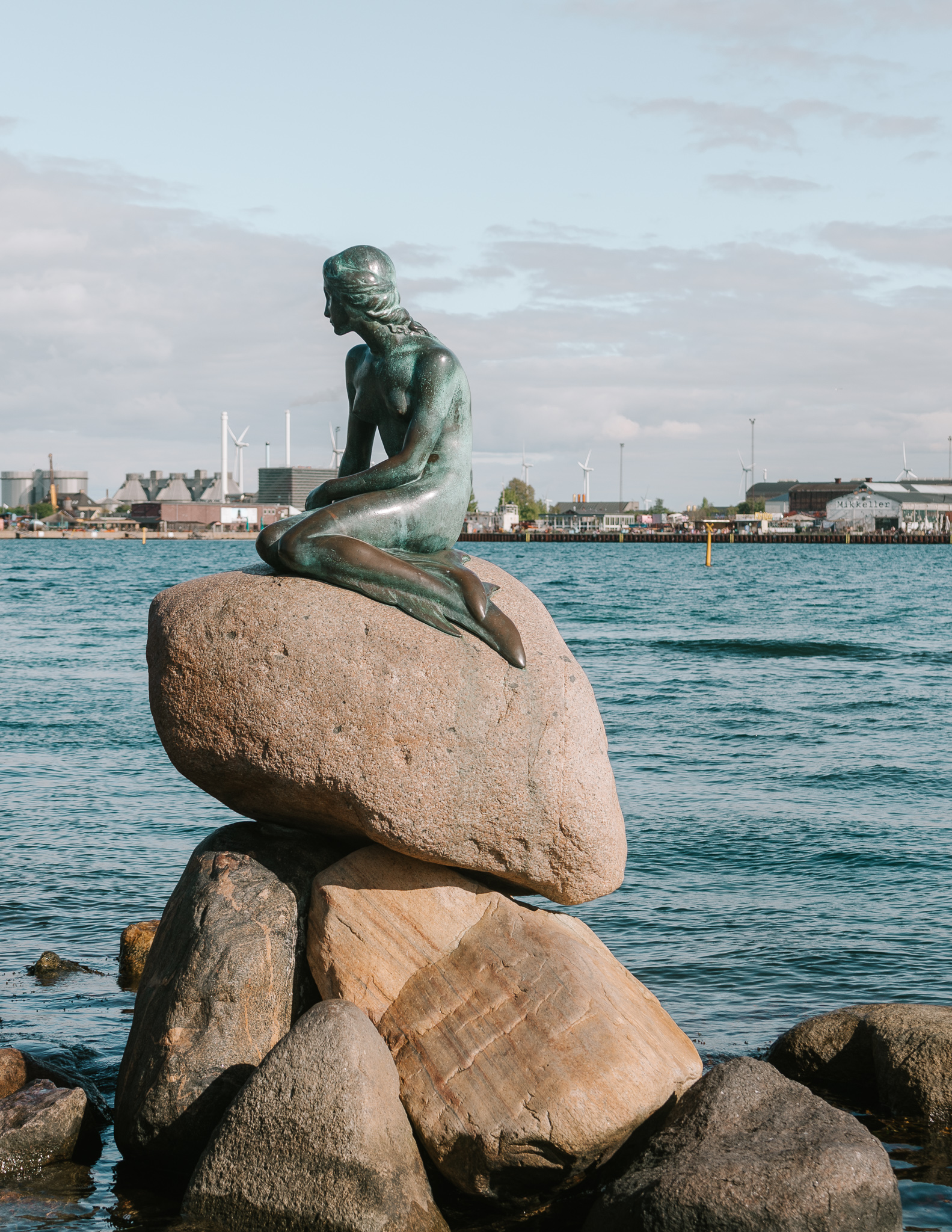 The little Mermaid stayue on a pile of large boulders stakced on eachother, next to a river with windmills on the banks behind in Kastellet, Copenhagen.