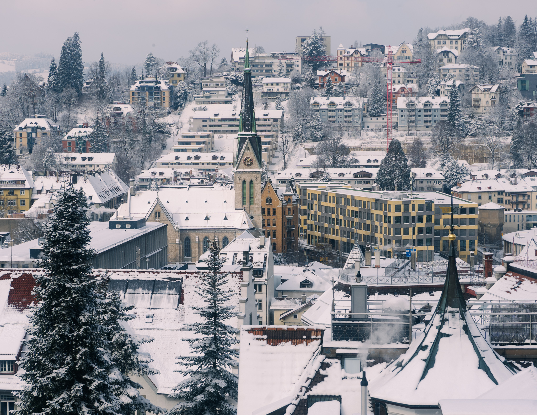 A view of st. gallen from above in winter, with snow on the church, buildings, trees and hills.
