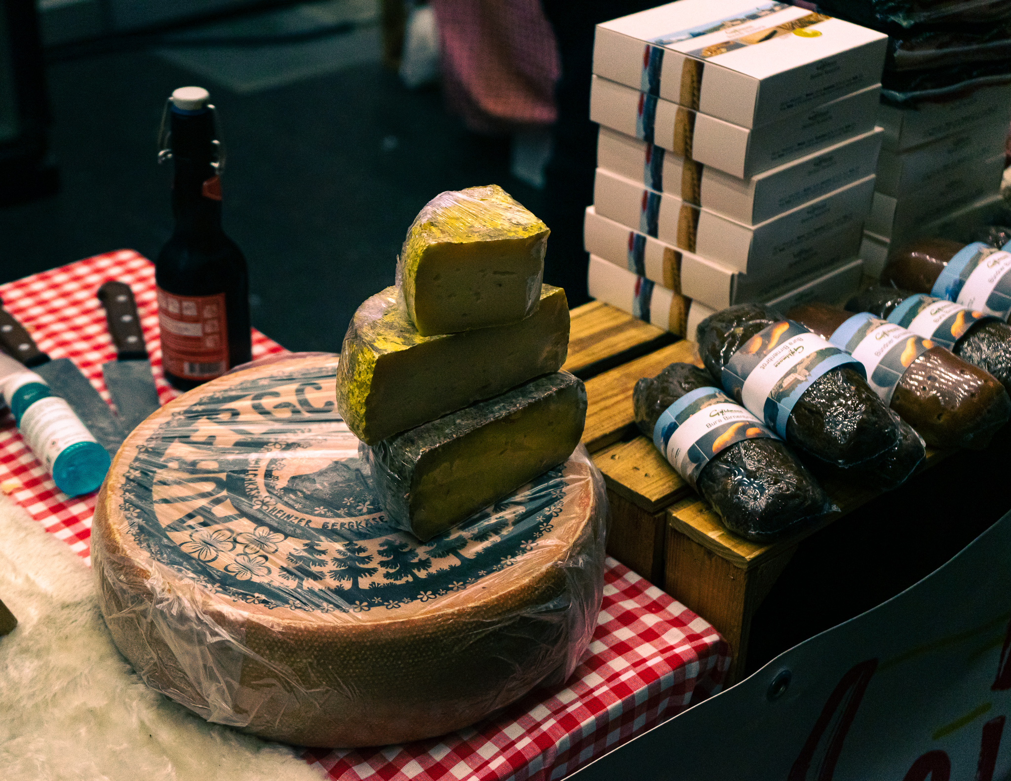 A wheel of Swiss cheese, with a stack of cut cheese on top, packed sausages to the right, and a bottle of cider behind, on a checkered cloth.