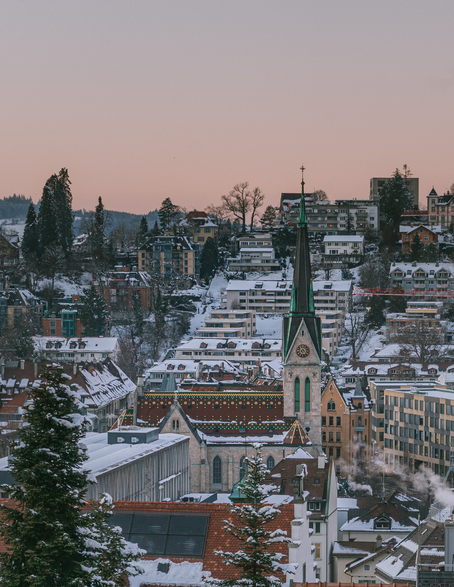 A pink sunset views of St. Gallen in winter, with the church in the center, and surrounding buildings and trees covere in snow.