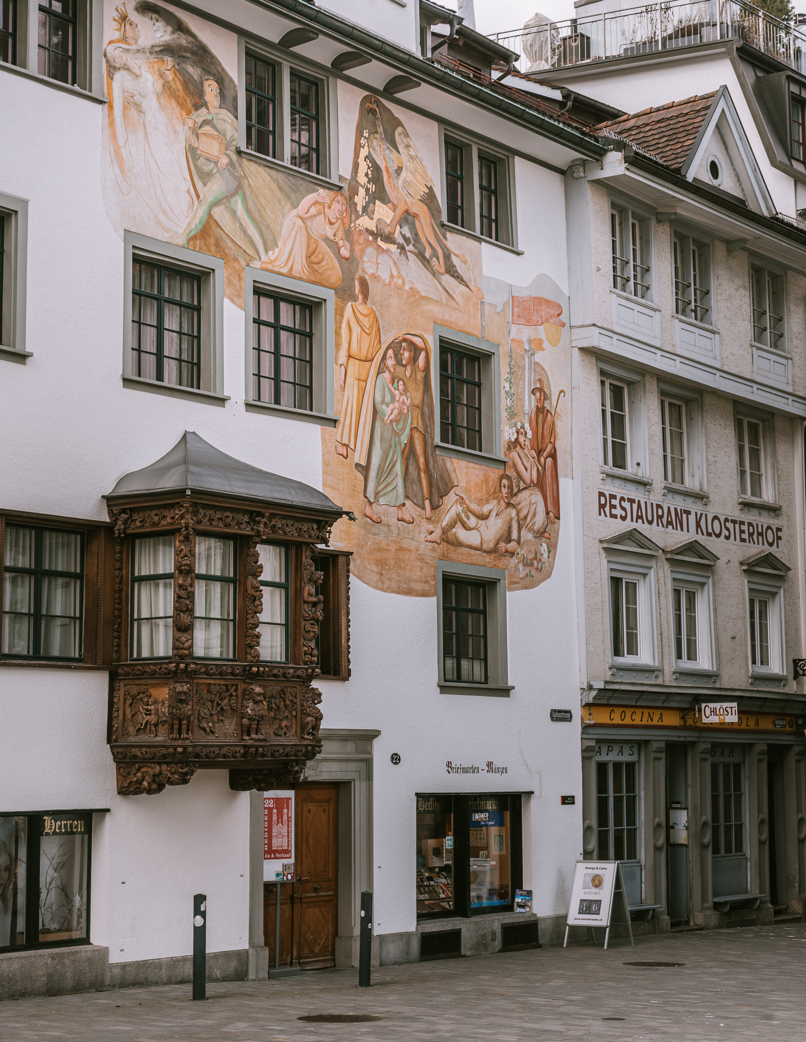 A facade of a house in St. Gallen, featuring a detailed, carved, wooden window and a colorful fresco of some peasant farmers and what looks to be Mary, Joseph and Jesus.