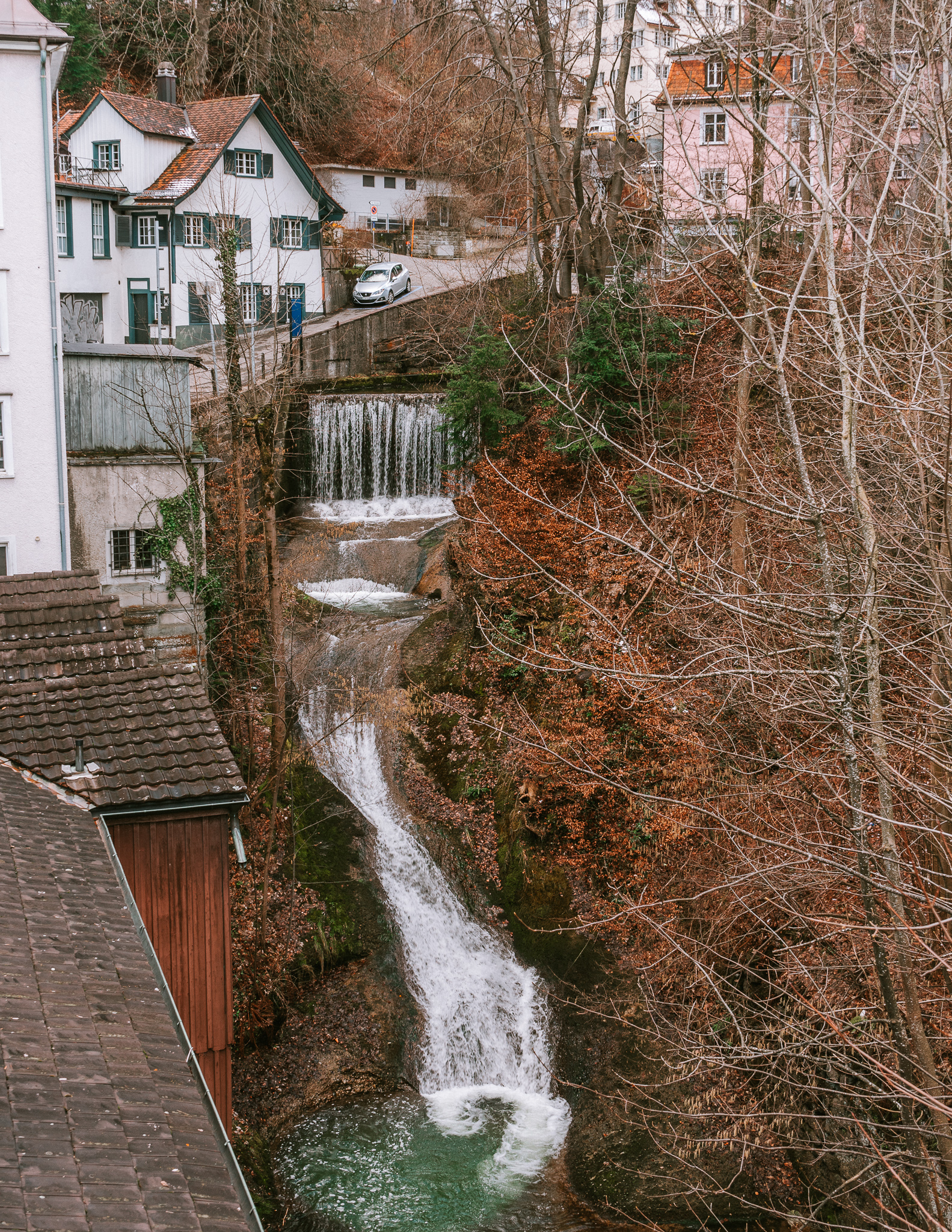 A waterfall running through the hills if St. Gallen, with houses on either sides, and trees with orange and green leaves.