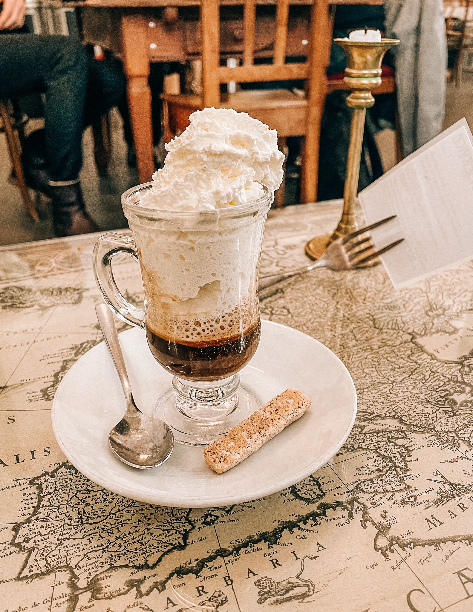 A glass cup of hot coffee with a heaping pile of whipped cream on top, on a table featuring a picture of a vintage map. 