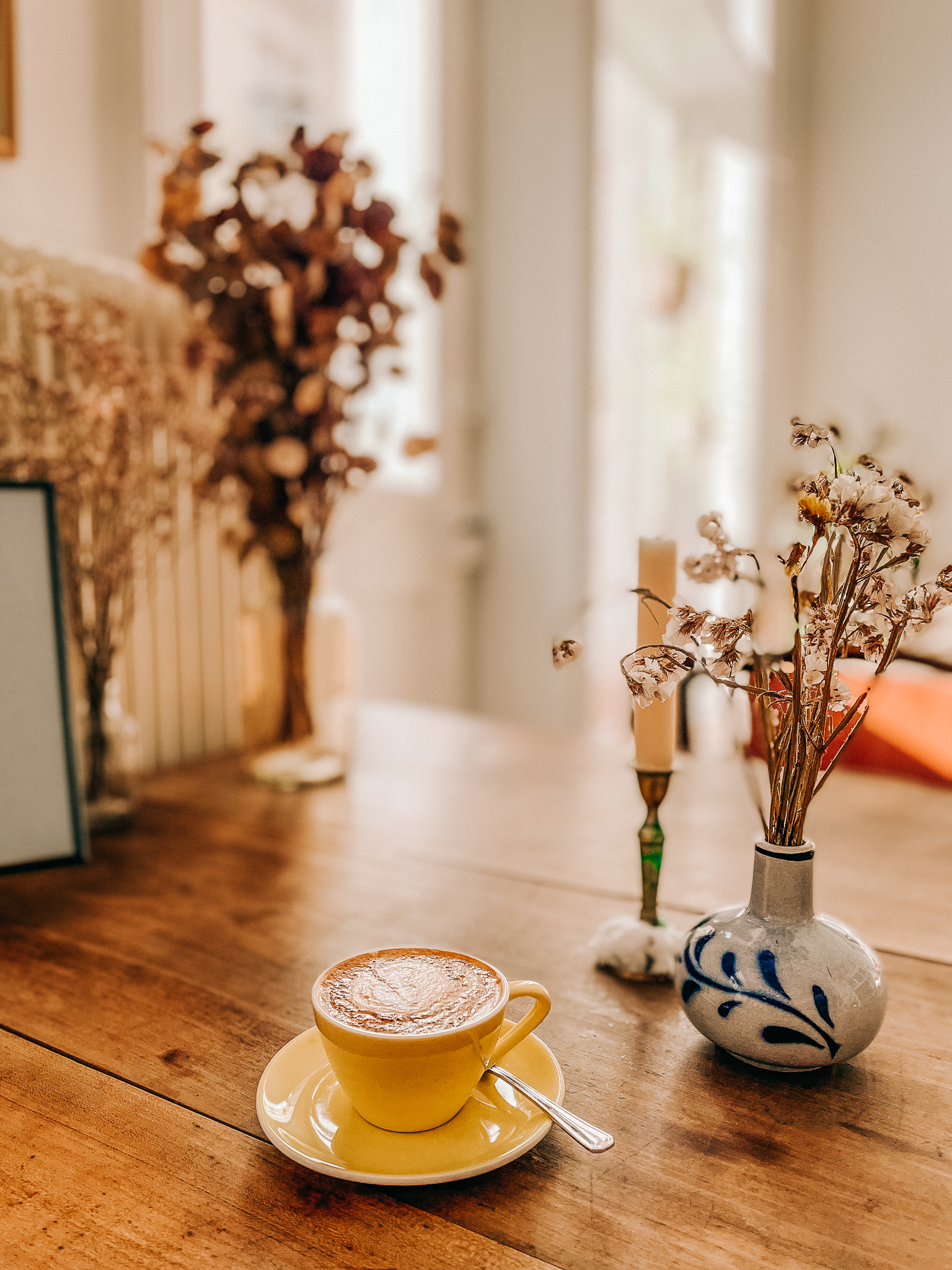 A coffee, vase of dried flowers, and a candle on a wooden table on a sunny day in Otmar Cafe in St. Gallen