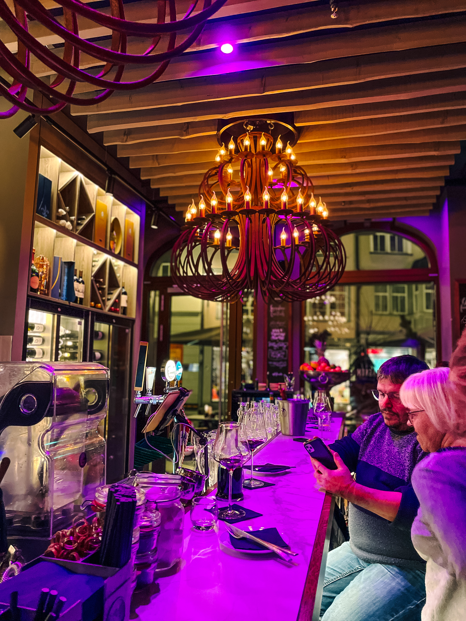 A view of the Magnum Wine Bar's bar seating area at night, featuring an elaborate iron cast chandelier with candles, purple low-lighting and people sitting at the bar. 