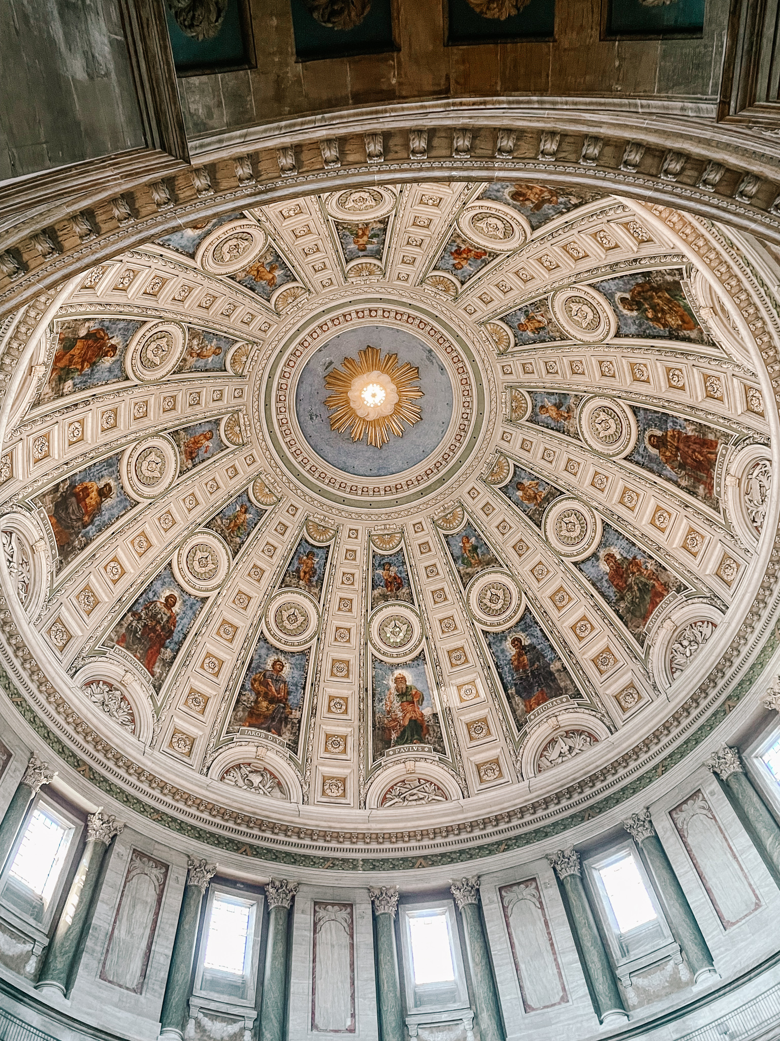 The domed roof of the Marble church in Copenhagen. It has a sun design in the center with rows of frescoes and ornate white marble designs radiating from the middle to the outside of the dome.