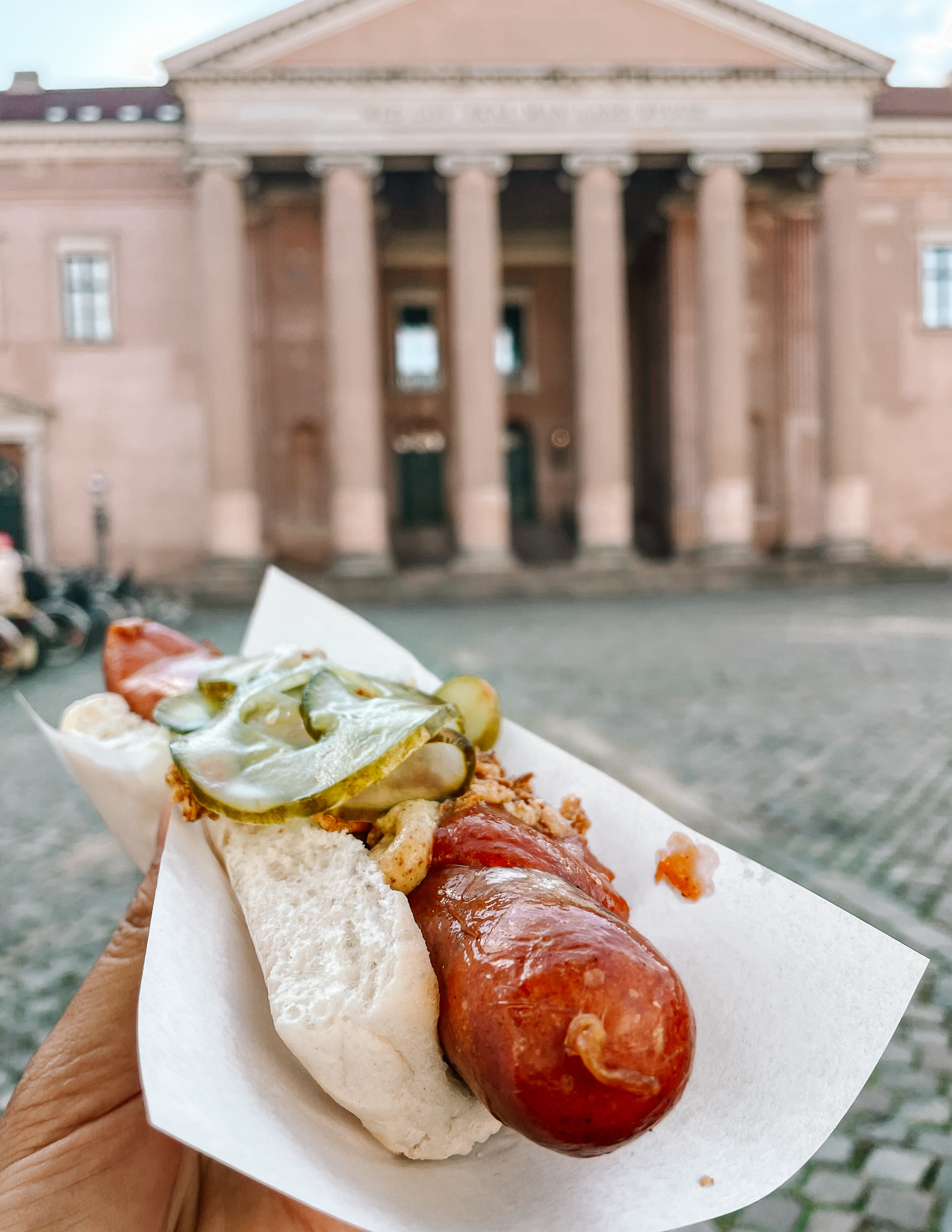 A Danish style hotdog with fried onions and pickles in Copenhagen in front of a historic roman style building