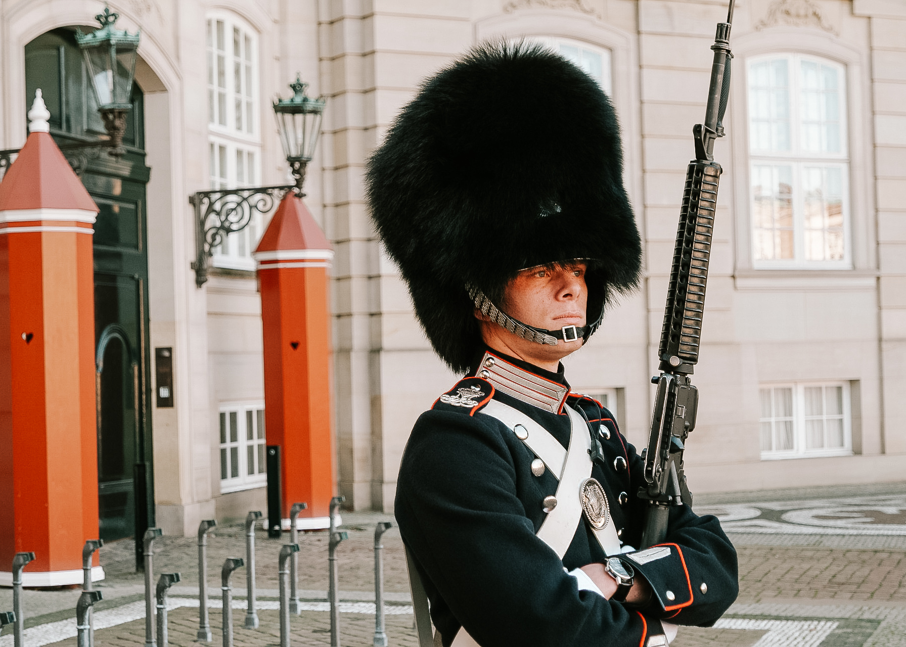 A guard in front of Amalieborg palace in Copenhagen with a fussy hat, a rifle and a navy traditional uniform in front of two red guard posts.