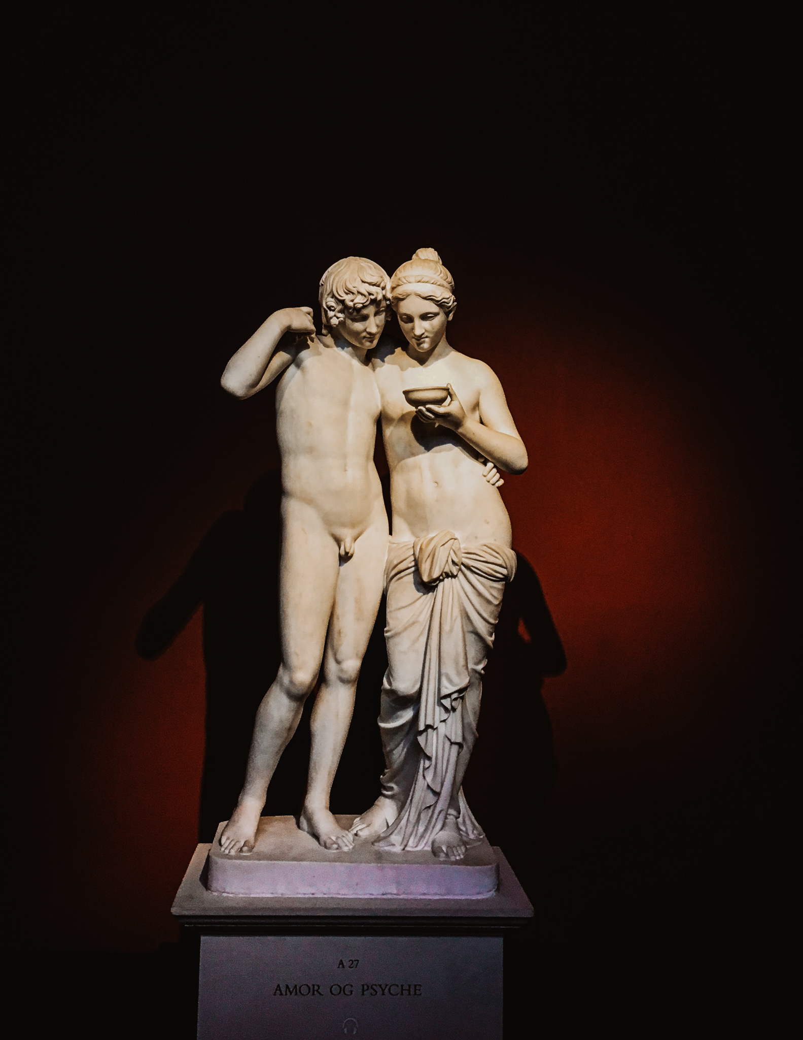 A marble statue of two human figures,1 man and 1 woman, in a dark room with one light source.
