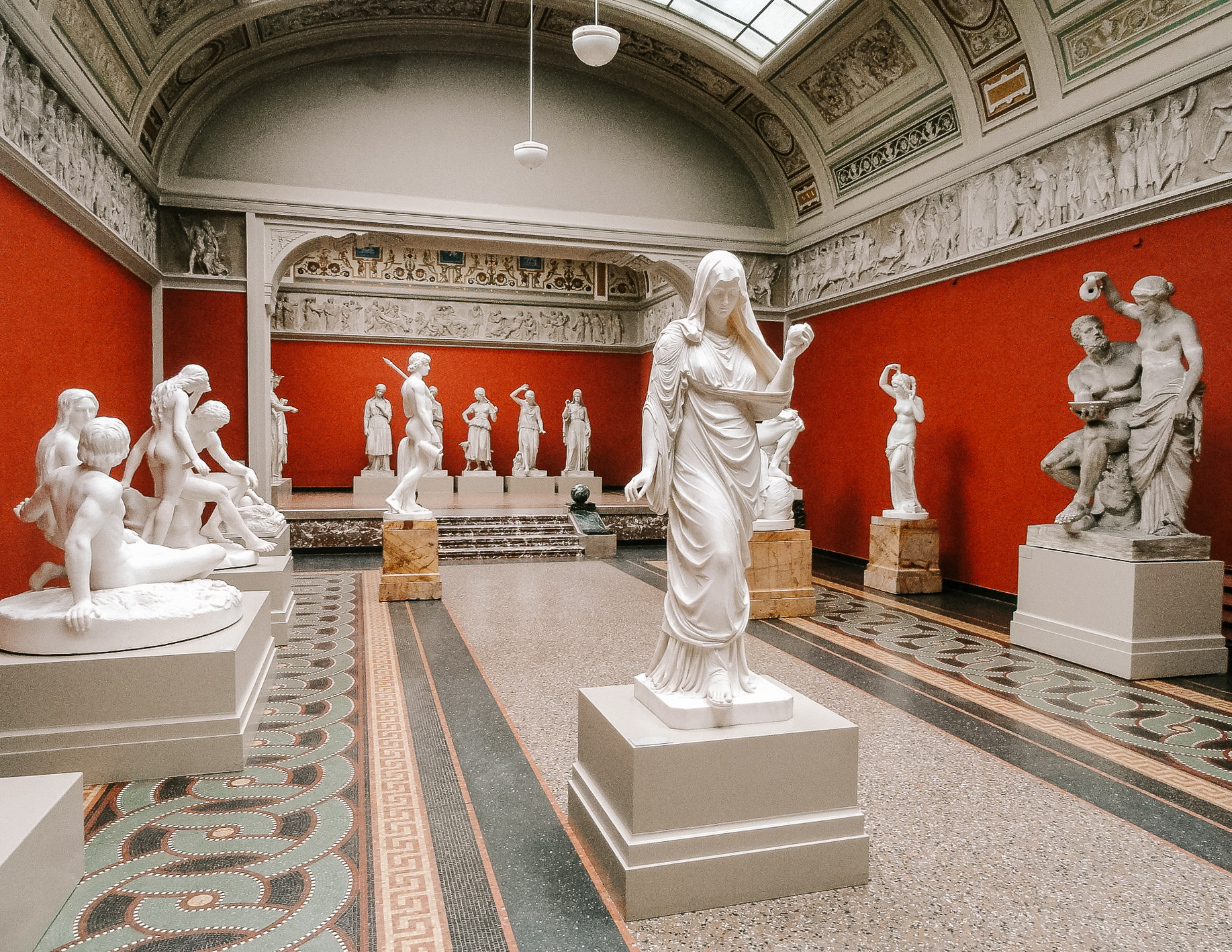 The interiors of a museum in Copenhagen in with many white marble sculptures of human figures in a red room.