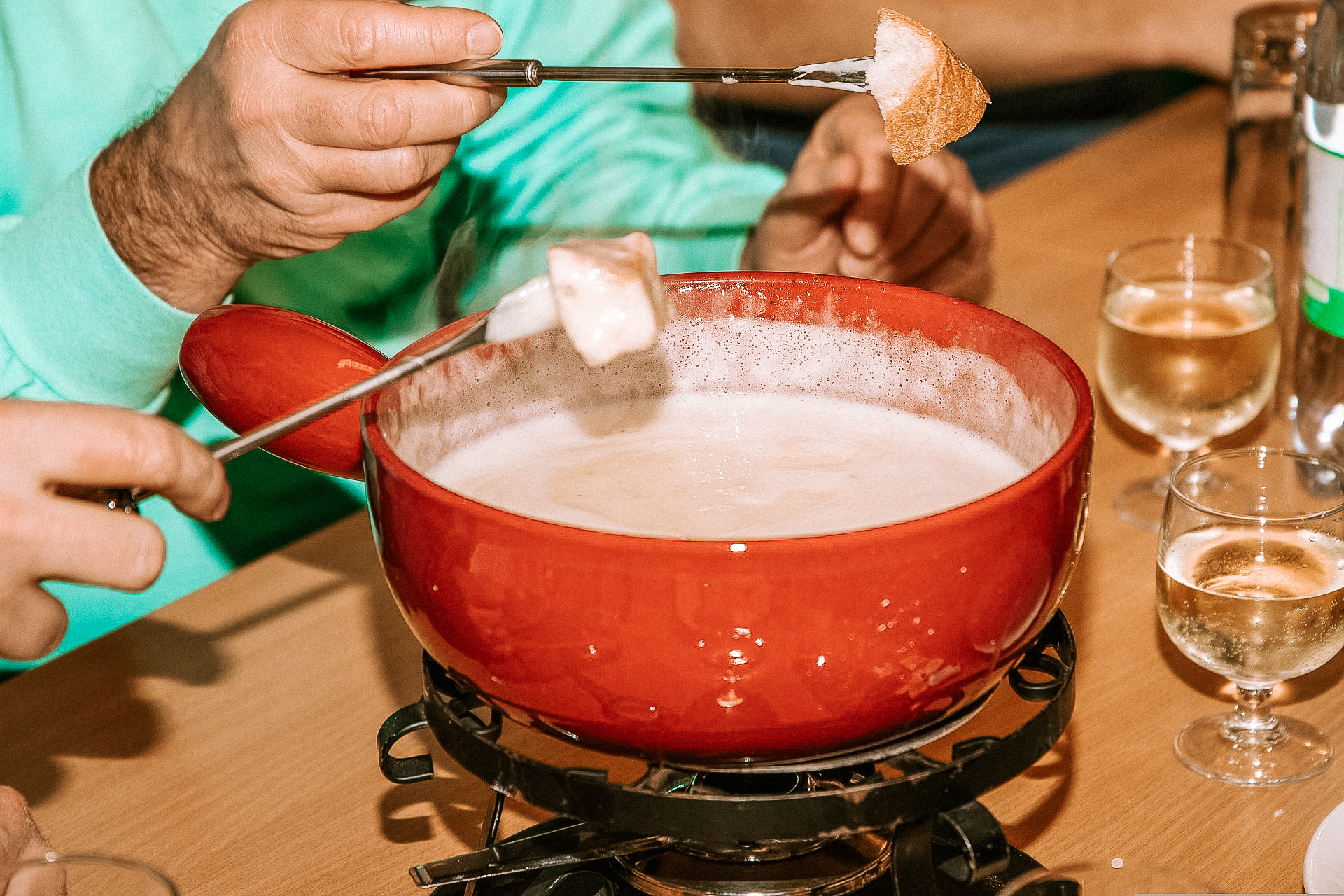 Two people enjoying a pot of fondue with bread on some prongs, dipped in the cheese, in a red pot, with two glasses of white wine on the side.