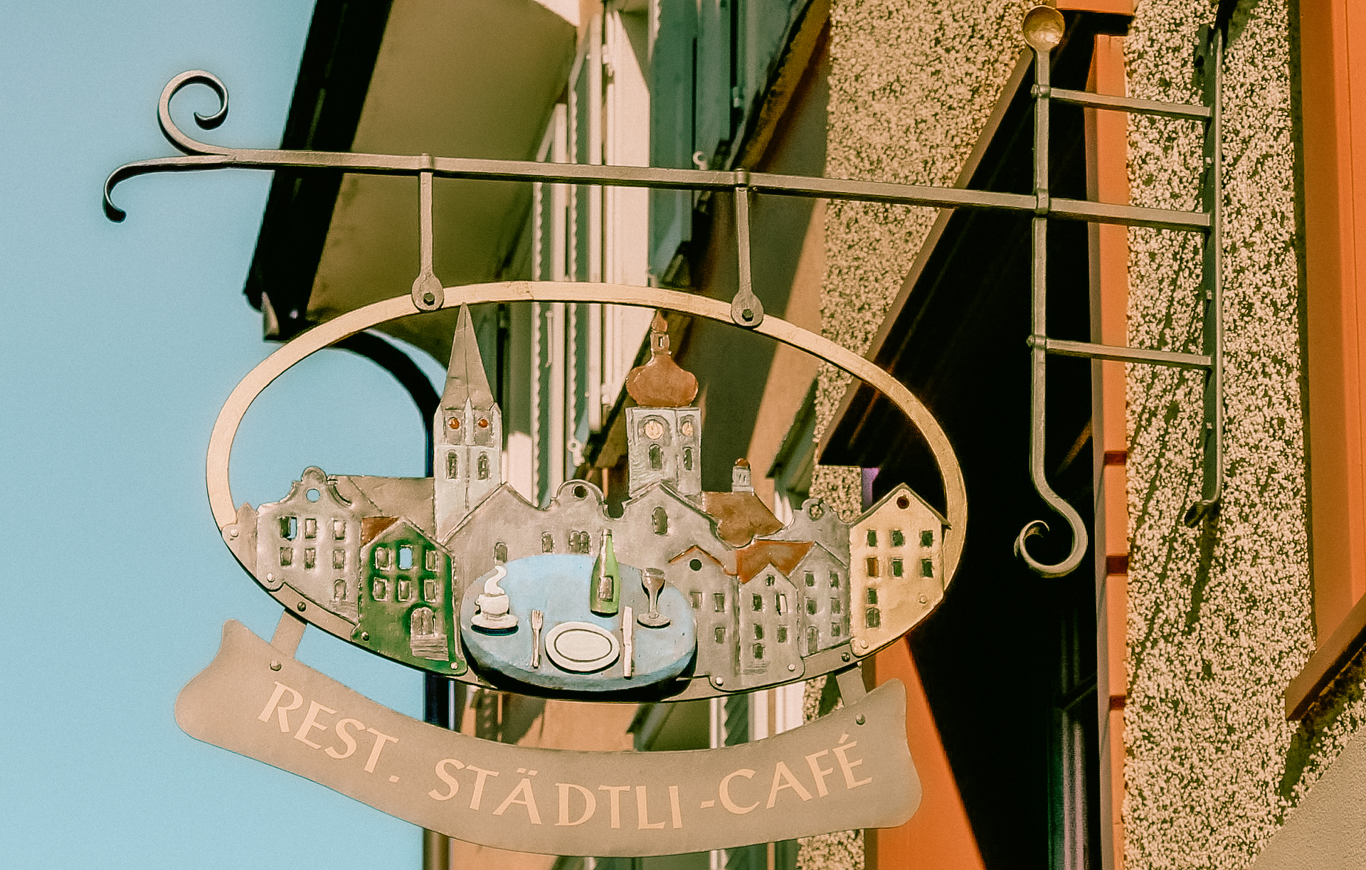 an old, colorful shop sign in st. gallen for a restaurant cafe with a small version of St. Gallen, a bottle of wine, a plate and a glass of wine on it.