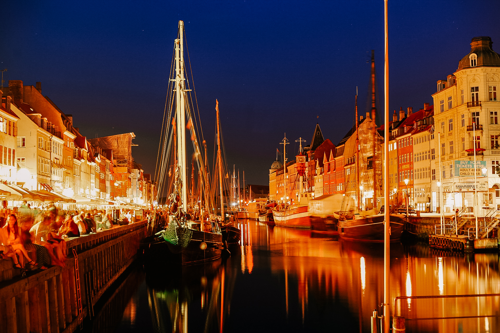 A pictre of Nyhavn canal in Copenhagen at night, with pictures of boats and well lit up restaurants lining the sides.