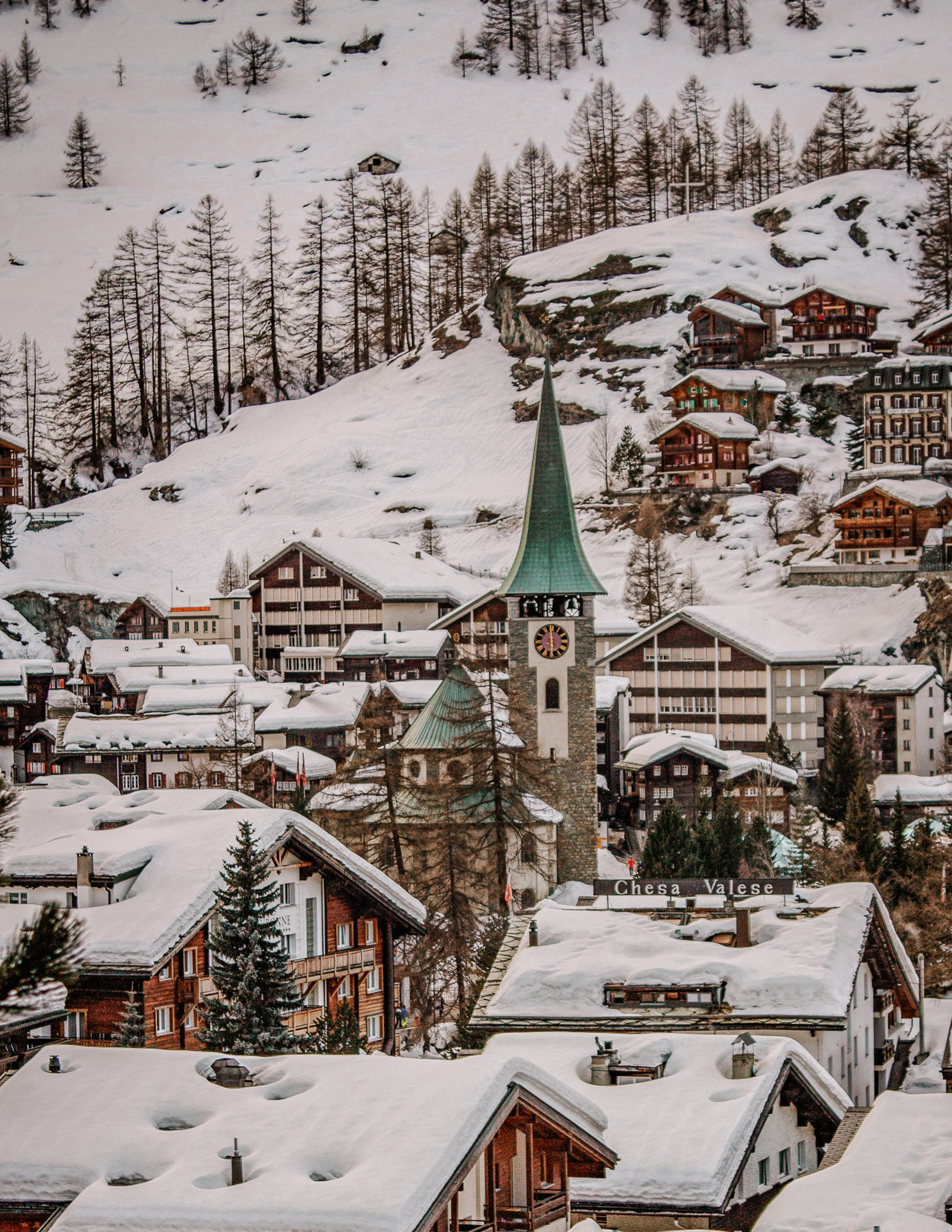 View of Zermatt during Christmas from above with Zermatt church and surrounding wooden chalets covered in snow, with bare trees on the mountain beyond.