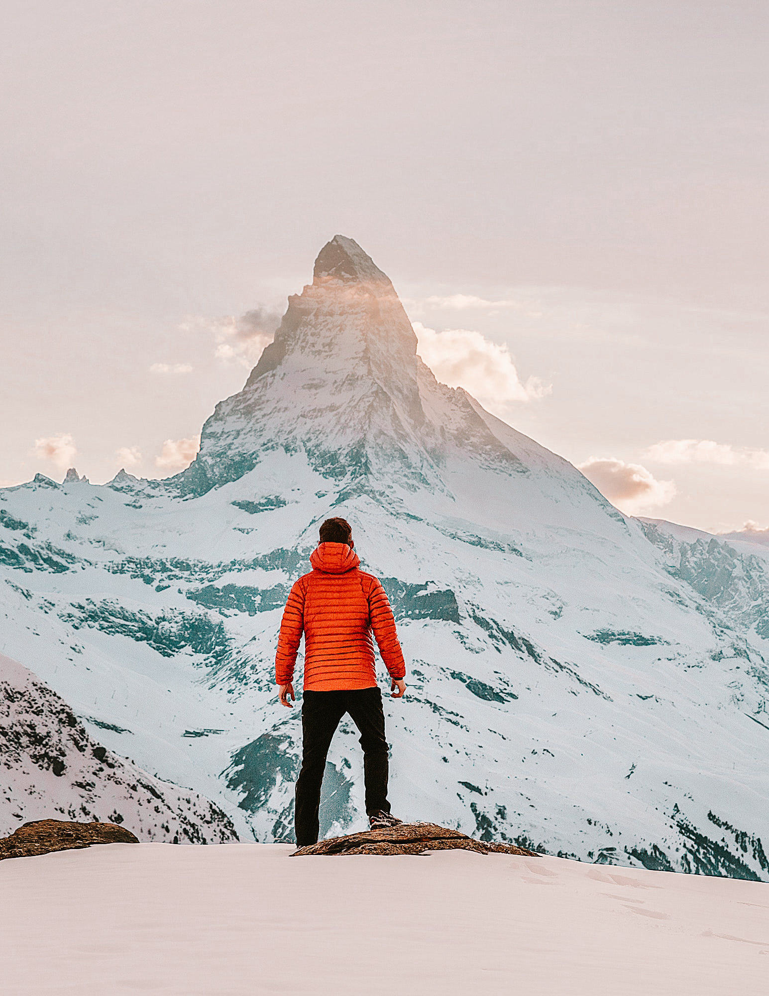 A man in a red jacket standing in front of the Matterhorn near Zermatt in winter. There is snow everywhere and soft sunlight.