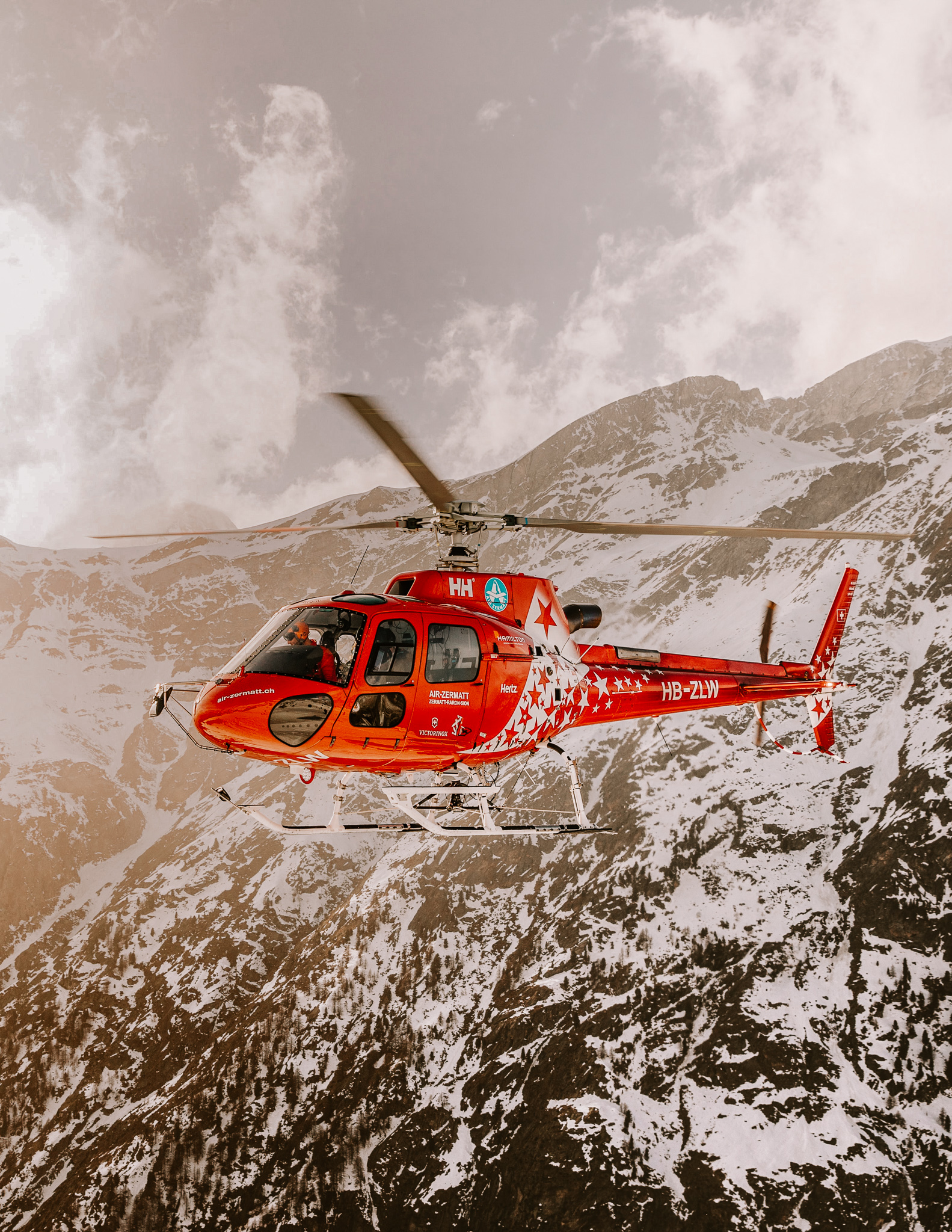 A single red helicopter suspended in middair, in front of a snowy mountain in the alps