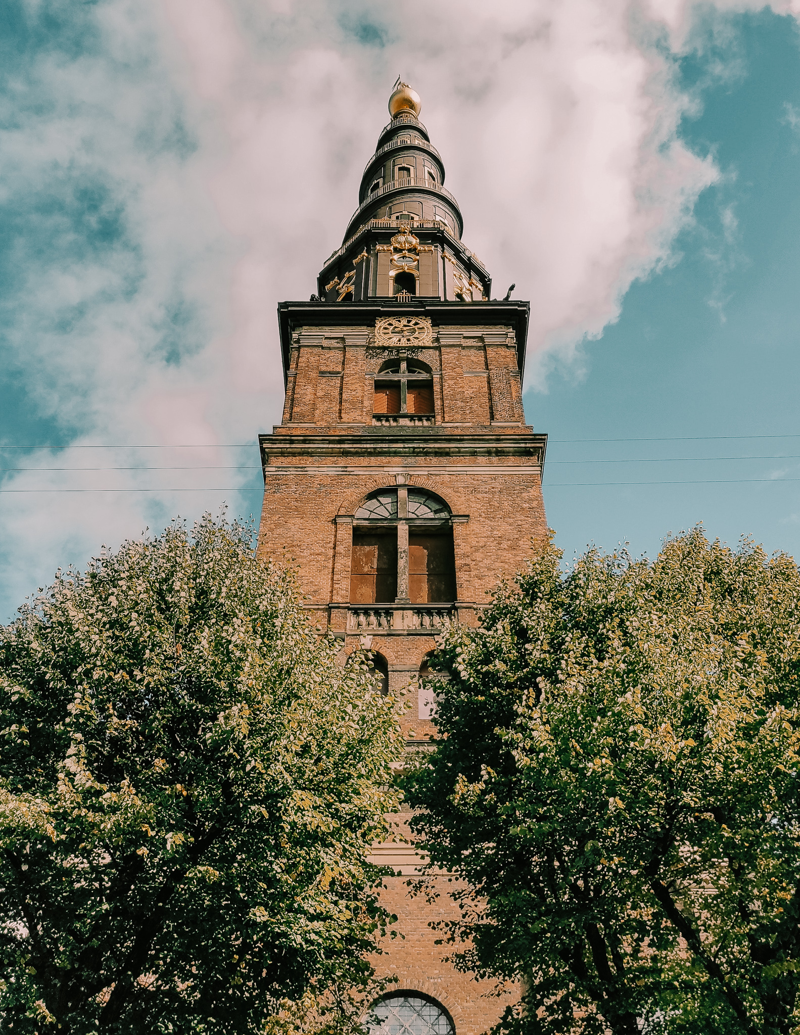 The tower of The Chruch of our Savior in Christianshavn, Copenhagen. It has brick throughout the exterior with a brown spiraled tip with a golden ball at the top of the tower.