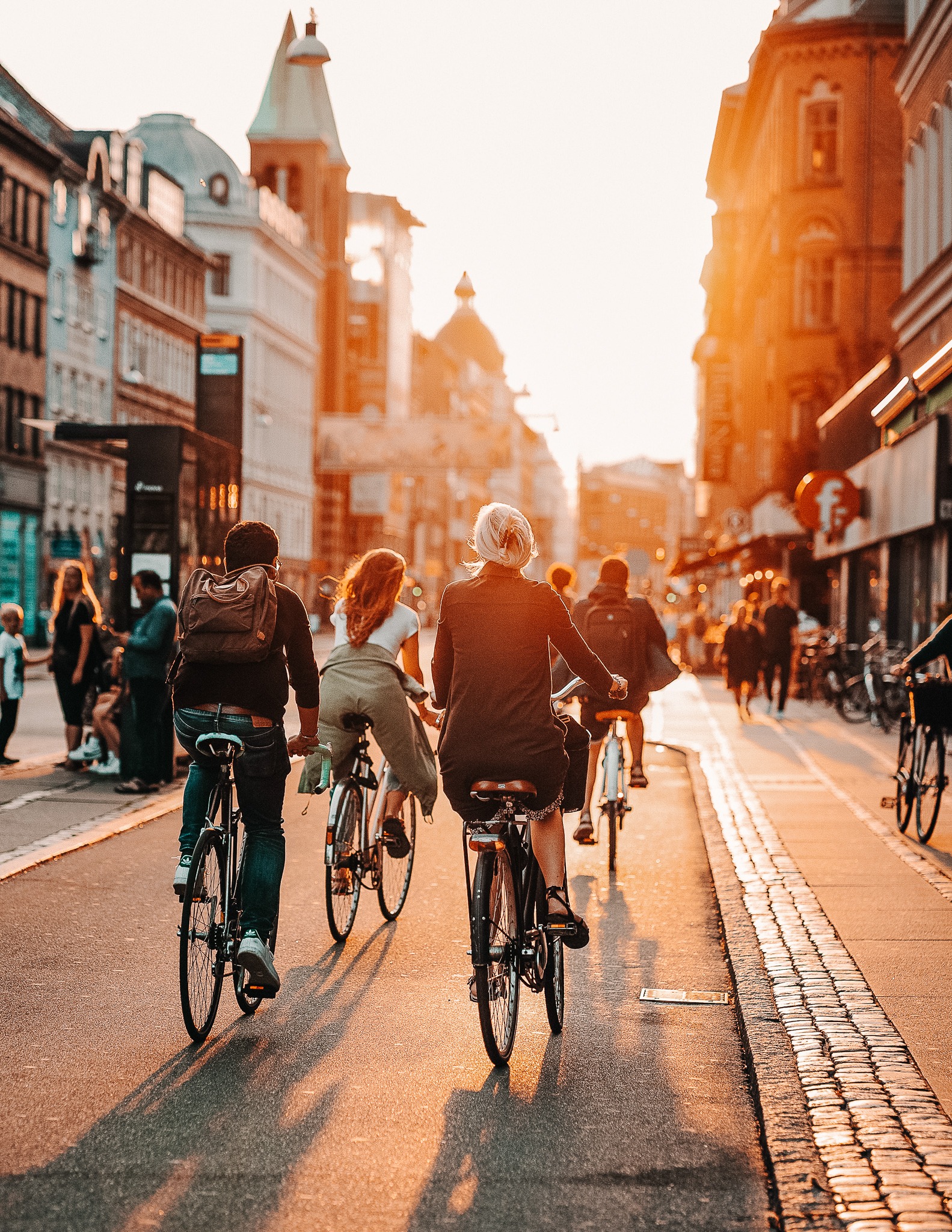 3 cyclists biking in a bike lane in Copenhagen, with pedestrians in the background during sunset.