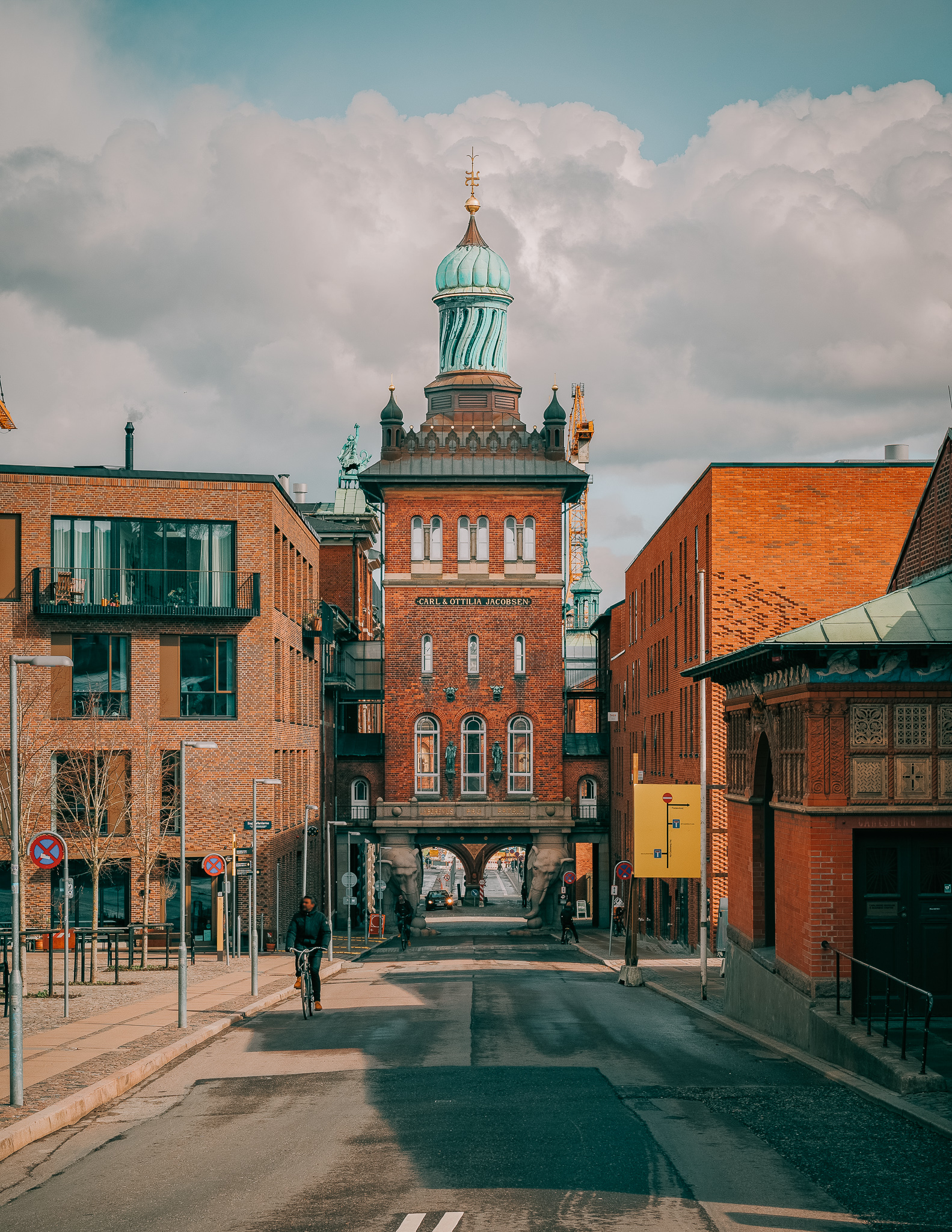 A towered brick building with a spiraled green copper tip, with arches going over a small street in Copenhagen