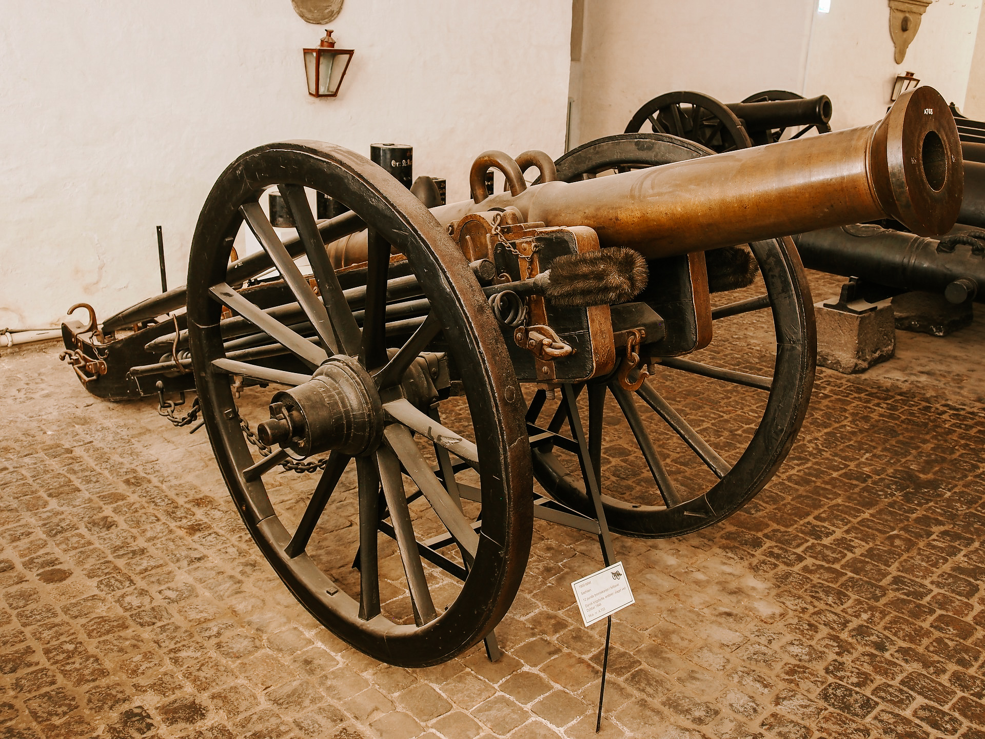 A picture of a metal canon that looks worn, in a white room with cobblestone floors.