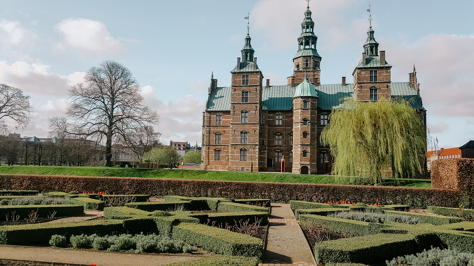 A view of Rosenburg castle in Copenhagen from the side, with a garden of trimmed hedge busges in the foreground, and willow trees near the castle.