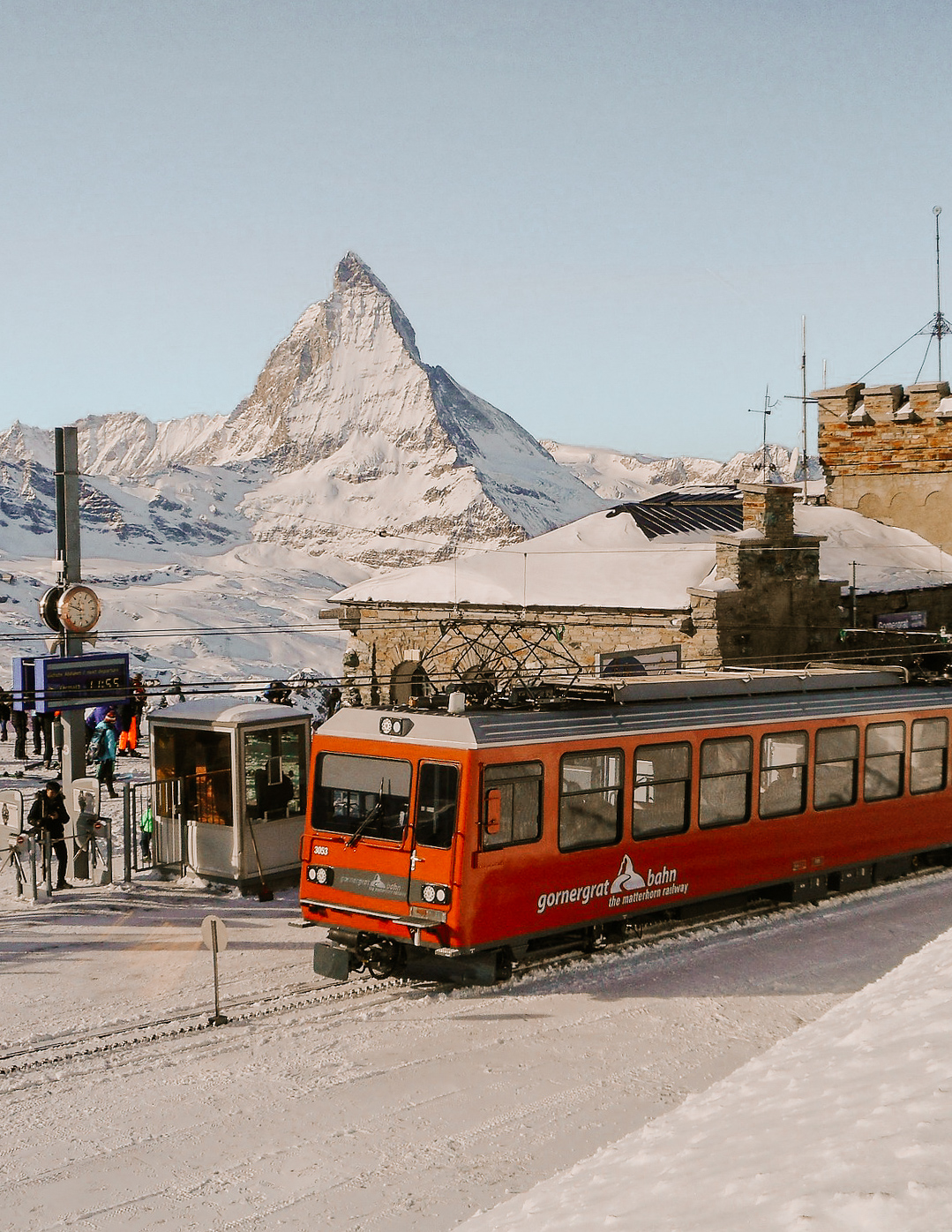A red cogwheel railway train in front of the Matterhorn in winter, with several people standing nearby on the train platform.