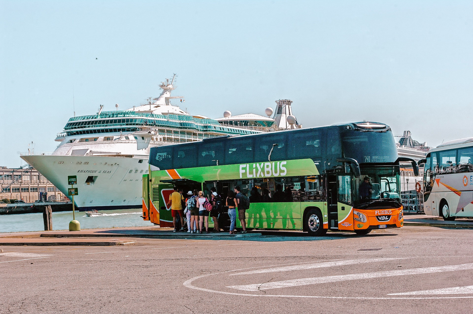 A picture of a green Flixbus in a parking location at the edge of a port with a ferry in the background.