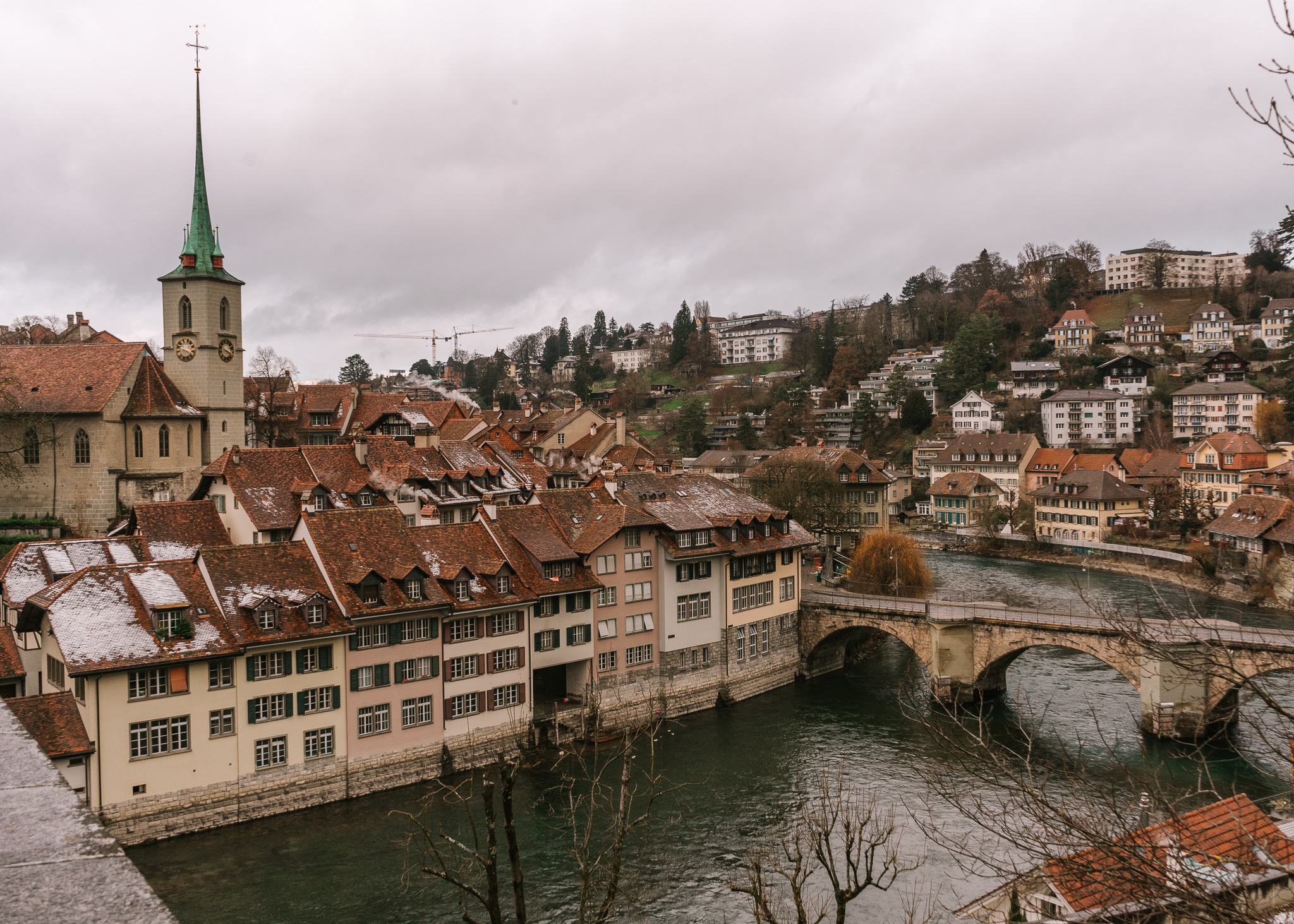 A view from above of the Old Town in Bern on a winter day. There is a bridge crossing the river into the town, with old homes lining the river front, and a single green church tower rising above.