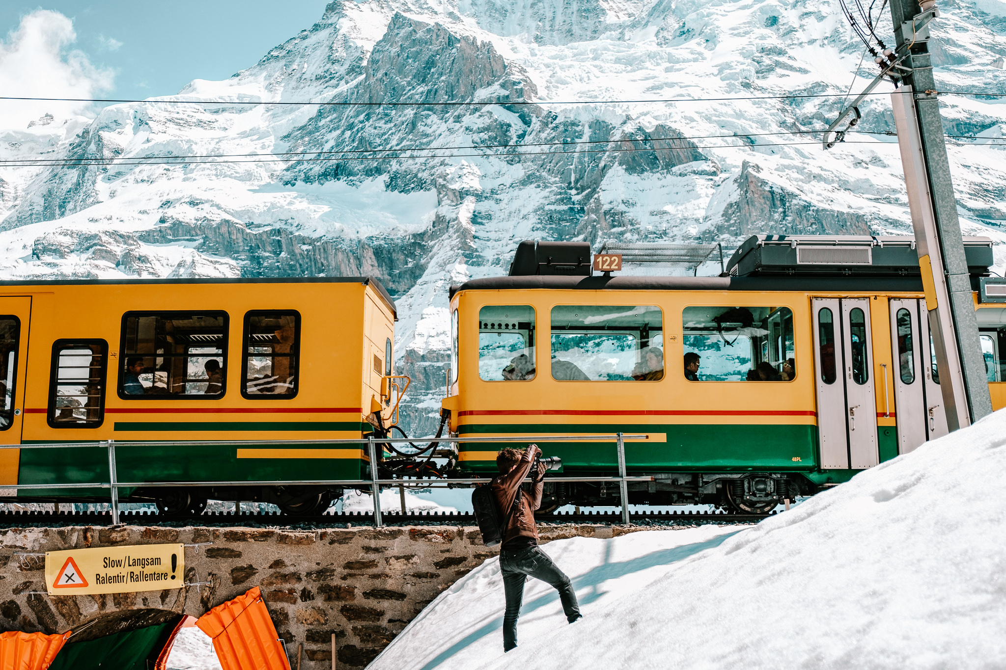 A yellow and green train running in front of a snowy mountain in the Swiss Alps, with a photographer taking a photo in front.