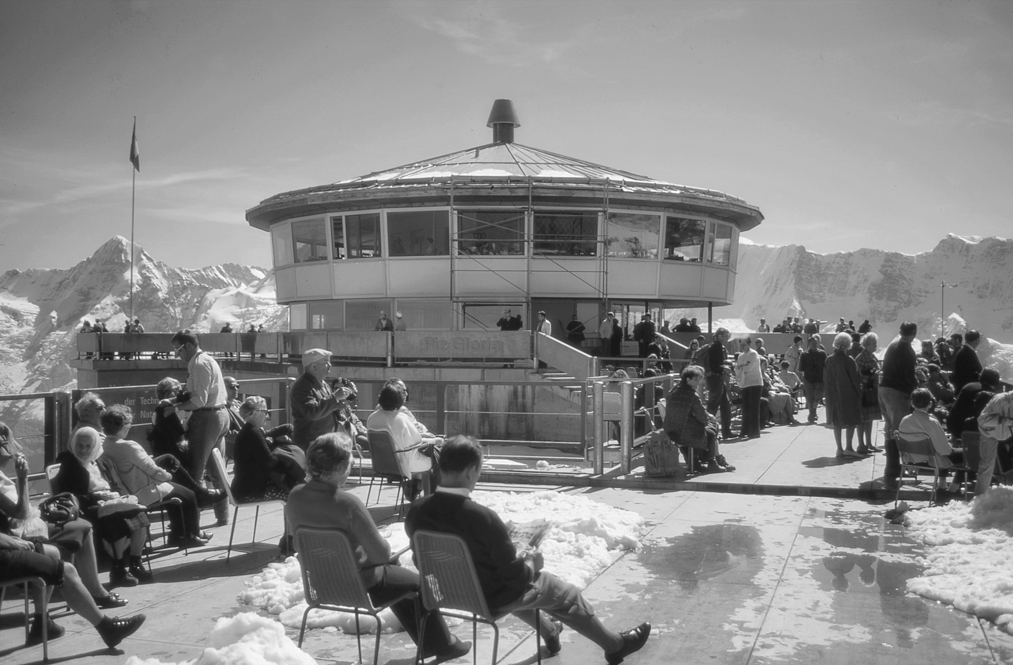 Many people sitting on a snowy terrace outside the circular Piz Gloria Restaurant during winter.
