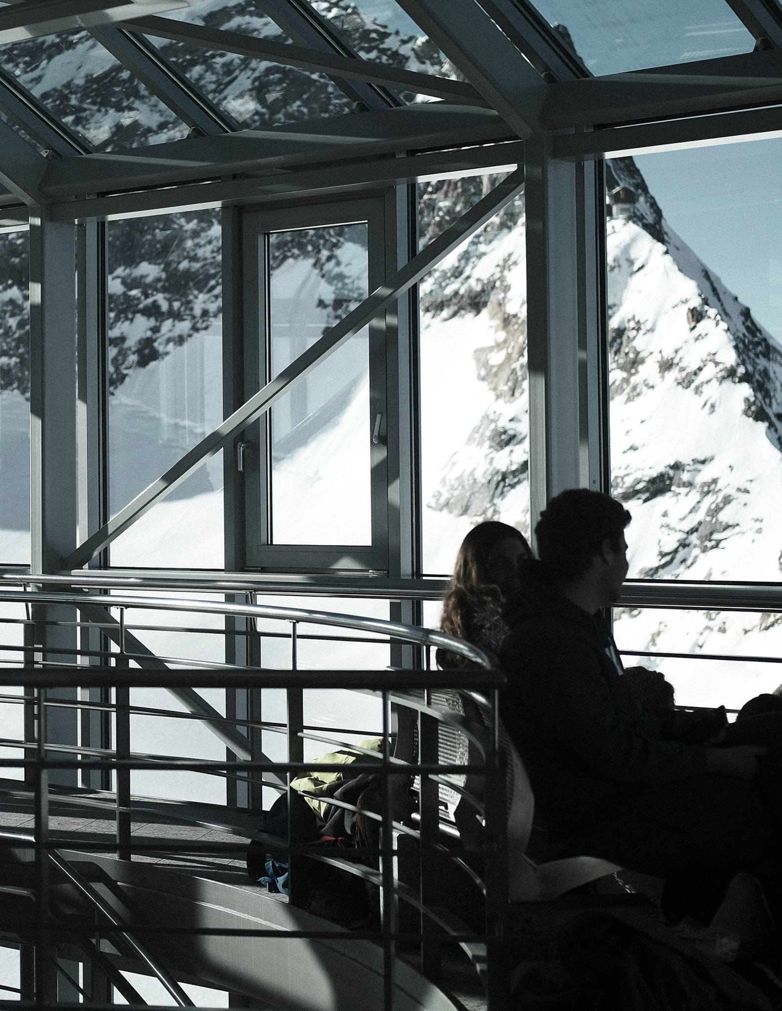 Two people sitting inside the Piz Gloria restaurant, made of large glass windows, with views onto the snowy Swiss Alps mountains just outside.