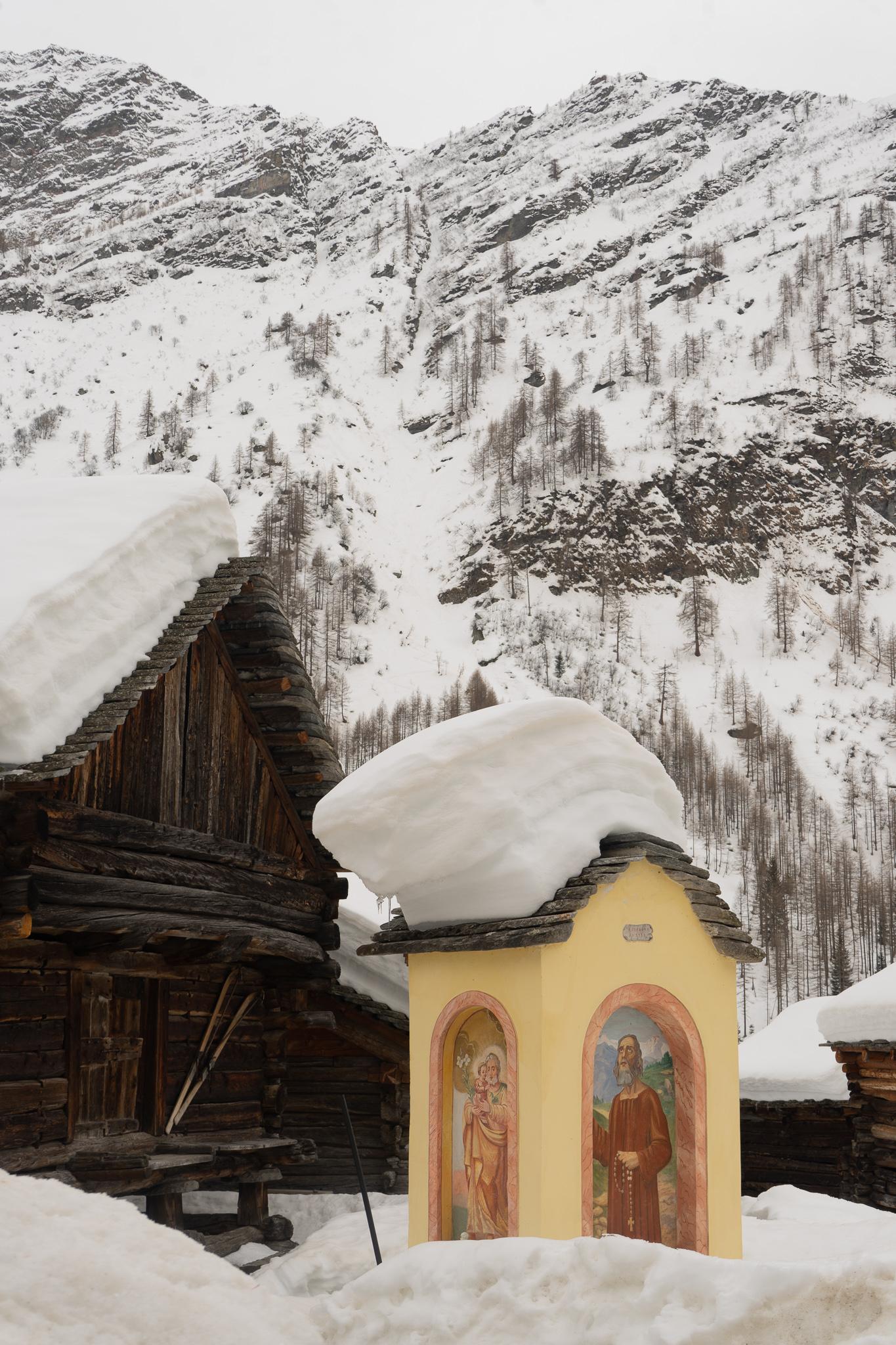 a snow covered mountain and wooden swiss chalet hut in front of it with heavy snow on its roof