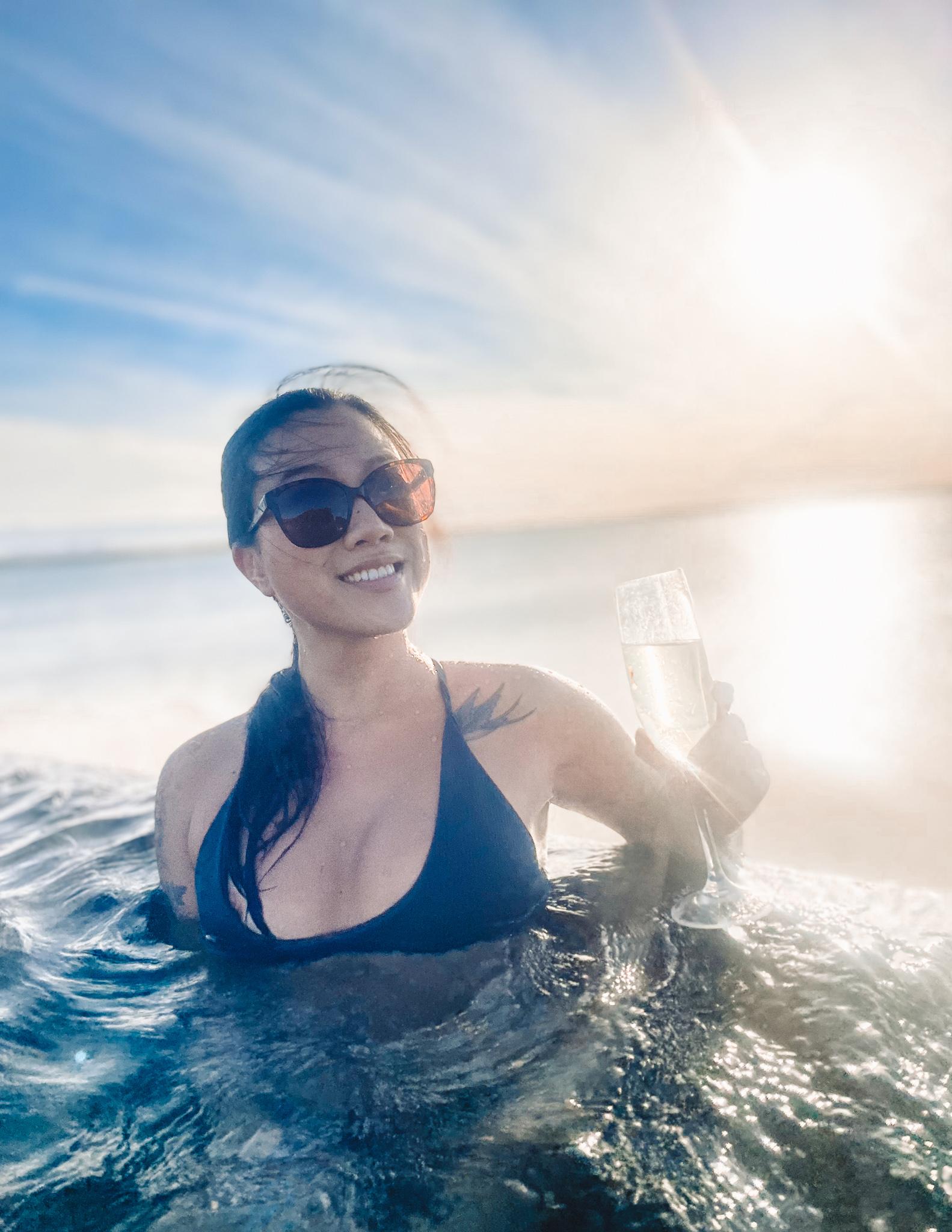 A woman smiling and holding champagne in an infinity pool overlooking the sea at the Sky Lagoon