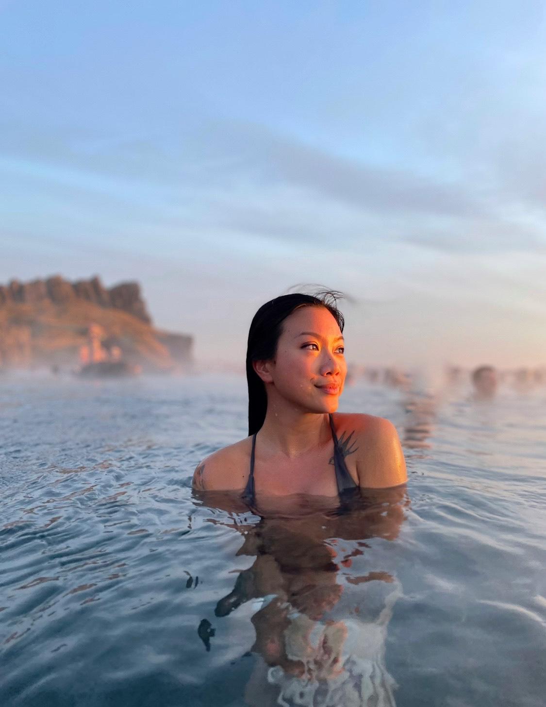 a woman in the Sky Lagoon geothermal bath during sunset in front of a tuft house sauna room in Iceland
